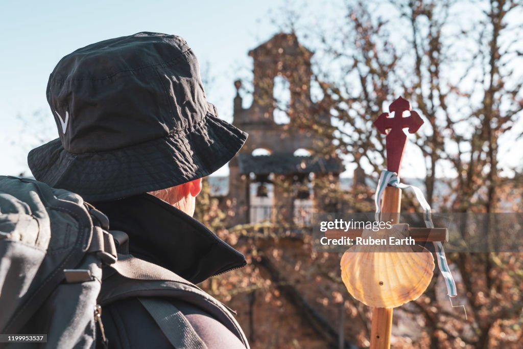 Santiago de Compostela way pilgrim with a hiking pole and shell looking at a church on the way.