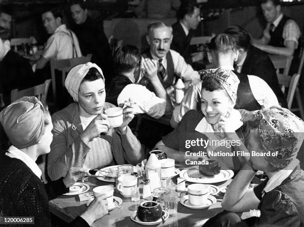 In the foreground, a group of four women, each wearing a working head scarf, are having lunch, smiling and discussing, while other male workers are...