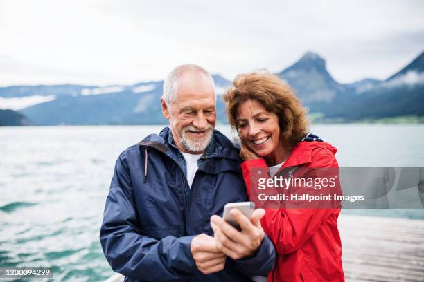 cheerful senior couple tourist standing by lake in nature on holiday, using smartphone. - mann zwei telefone stock-fotos und bilder