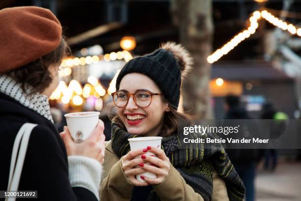 woman smiles at friend, holding coffee cup at christmas market. - christmas coffee stock-fotos und bilder