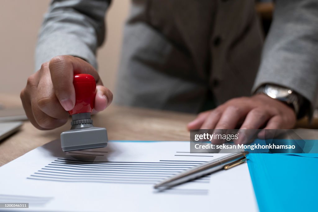 Close-up Of A Person's Hand Stamping With Approved Stamp On Document At Desk