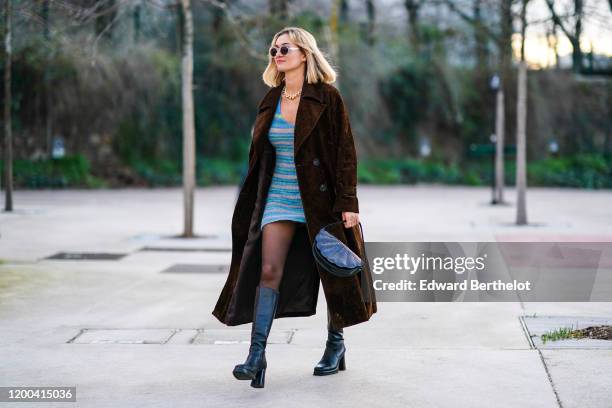 Anne-Laure Mais wears sunglasses, a brown long coat, a semi circular bag, black leather long boots, a blue striped dress, a necklace, outside...