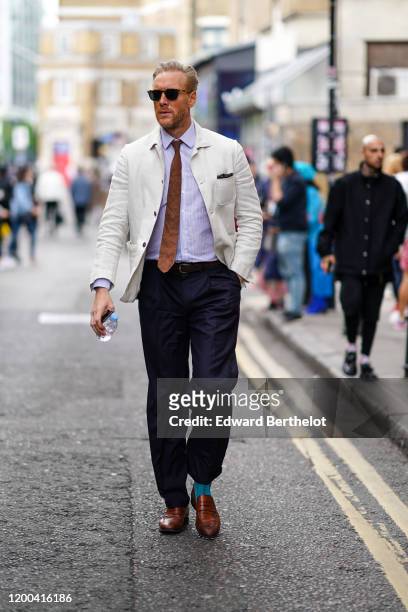 Guest wears sunglasses, a grey and white pinstriped shirt, a brown tie, a cream-color linen jacket, dark grey pants, turquoise blue socks, brown...