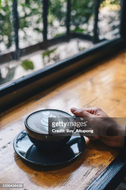 cropped shot view of someone hand touching a cup of goth latte or charcoal latte on wooden table. - goth boy stock-fotos und bilder