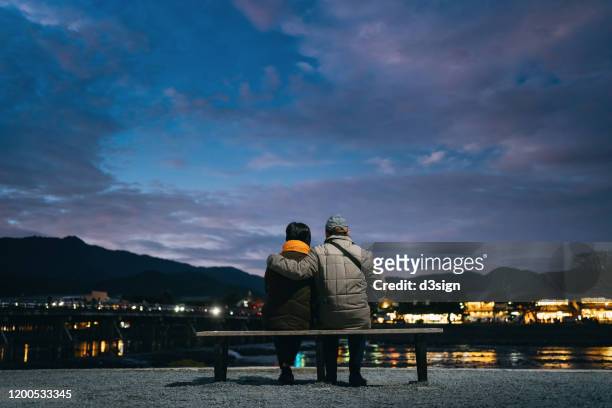 rear view of senior couple sitting side by side on a bench enjoying the sunset by the lake while visiting traditional japanese town in higashiyama, kyoto, japan at twilight - bench foto e immagini stock