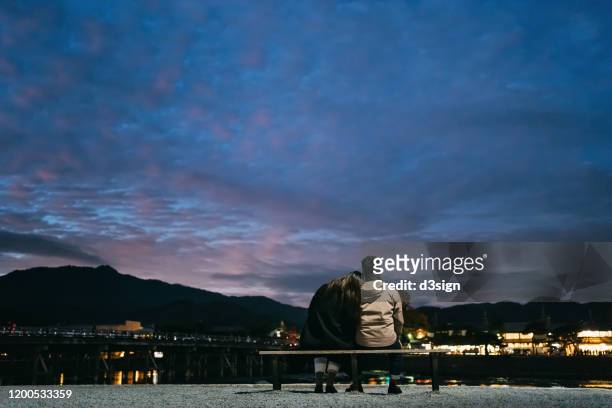 rear view of young couple sitting side by side on a bench enjoying the sunset by the lake while visiting traditional japanese town in higashiyama, kyoto, japan at twilight - first night of marriage stock pictures, royalty-free photos & images