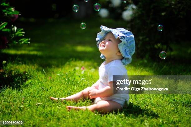 little baby girl in panama sits in the garden on the green grass. happy child smiles and plays with soap bubbles. outdoor games in summer - bubbles happy stockfoto's en -beelden