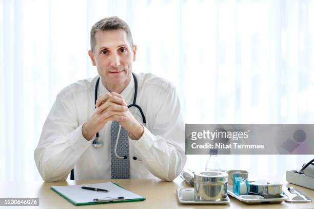 portrait of confident senior doctor wearing white coat with stethoscope sitting inside in office. professional occupation concept. - general practitioner imagens e fotografias de stock
