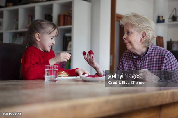 happy girl (6-7) putting raspberries onto grandmother's fingers at a dining table - child eat side photos et images de collection