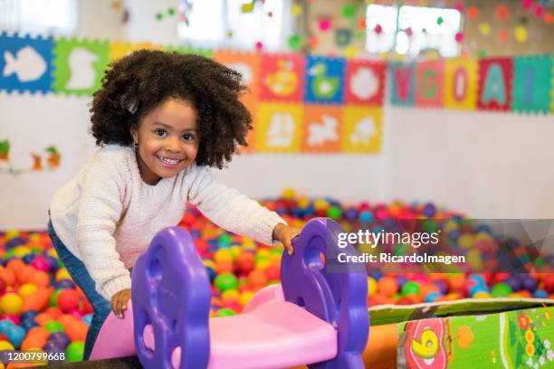 afro-haired and dark-skinned girl about four years old climbs up a pink and purple slide placed in a pool of colored balls while looking at the acamara and shows a beautiful smile - adult ball pit stock pictures, royalty-free photos & images