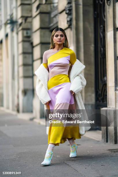 Thassia Naves wears earrings, a yellow and mauve striped flowing dress with mauve mesh inserts, a fluffy white coat, glittering sequined colorful...
