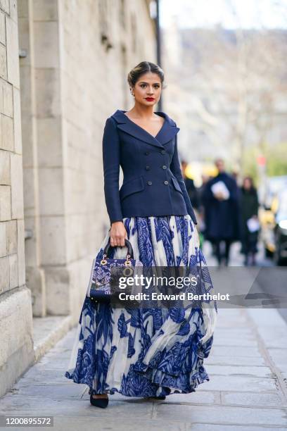 Thassia Naves wears earrings, a navy-blue v-neck jacket, a white and blue print long pleated skirt, a Dior embroidered bag, outside Dior, during...
