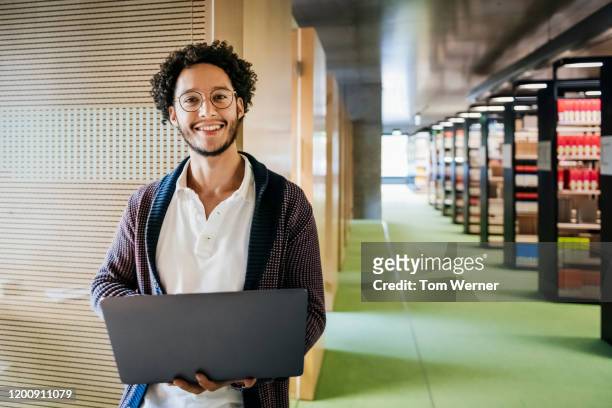 portrait of young man holding laptop in library - young adult fotografías e imágenes de stock
