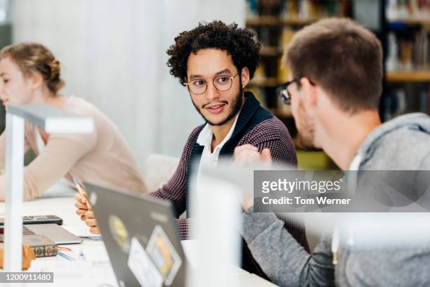 two students talking to each other while studying in library - male student wearing glasses with friends stockfoto's en -beelden