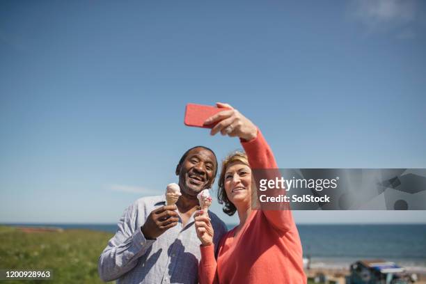 fröhliches senior-paar hält eis, das selfie am meer macht - eisstockschießen stock-fotos und bilder