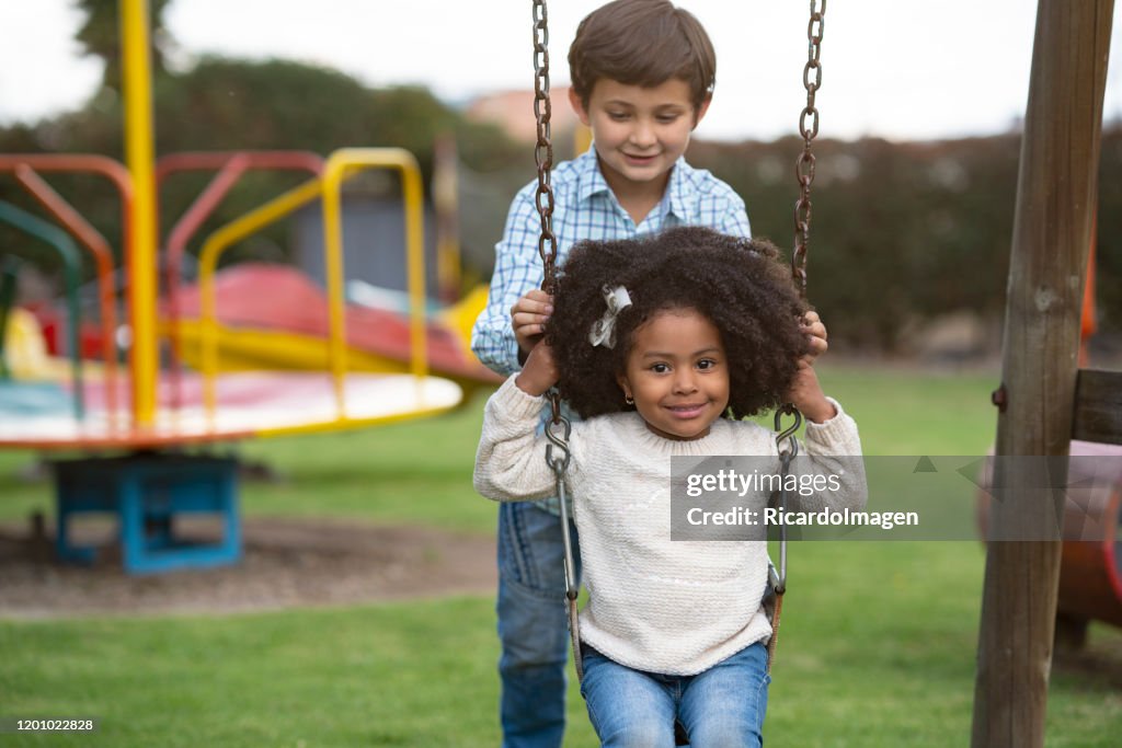 Latin boy with brown hair and approximate age of 8 years drives the swing in which his little Latin girl with afro hair and brown skin is sitting on a fun swing in a beautiful park