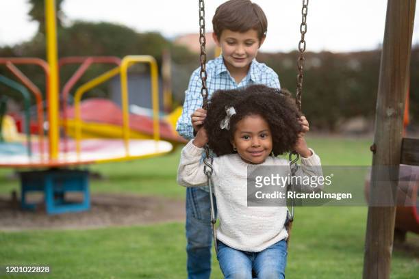 latin boy with brown hair and approximate age of 8 years drives the swing in which his little latin girl with afro hair and brown skin is sitting on a fun swing in a beautiful park - 8 9 years stock pictures, royalty-free photos & images