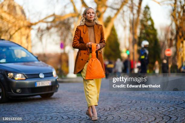 Leonie Hanne wears earrings, a brown knit turtleneck, a lustrous rust-color jacket, an orange woven leather bag, yellow leather crop pants, fishnet...