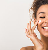Face care. Happy black woman applying moisturizer cream on cheek