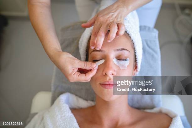 beautician applying patches under the eyes of a beautiful young woman - one eyed stock pictures, royalty-free photos & images