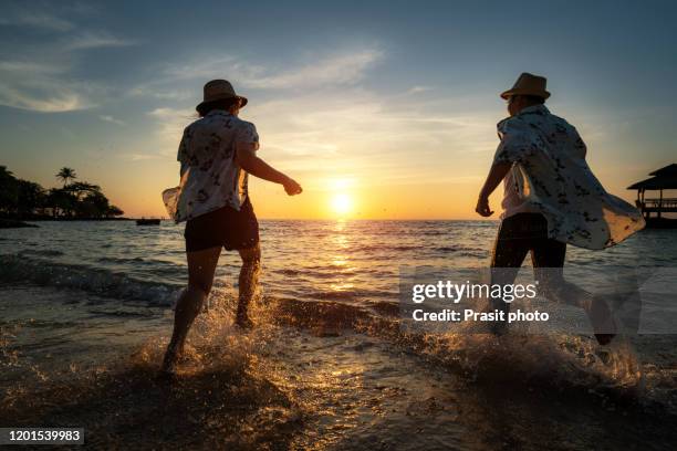 happy cheerful asian couple having fun running to the sea together and doing splashes of water on a tropical beach at sunset - concept about romantic vacation, honeymoon. - beach night stockfoto's en -beelden