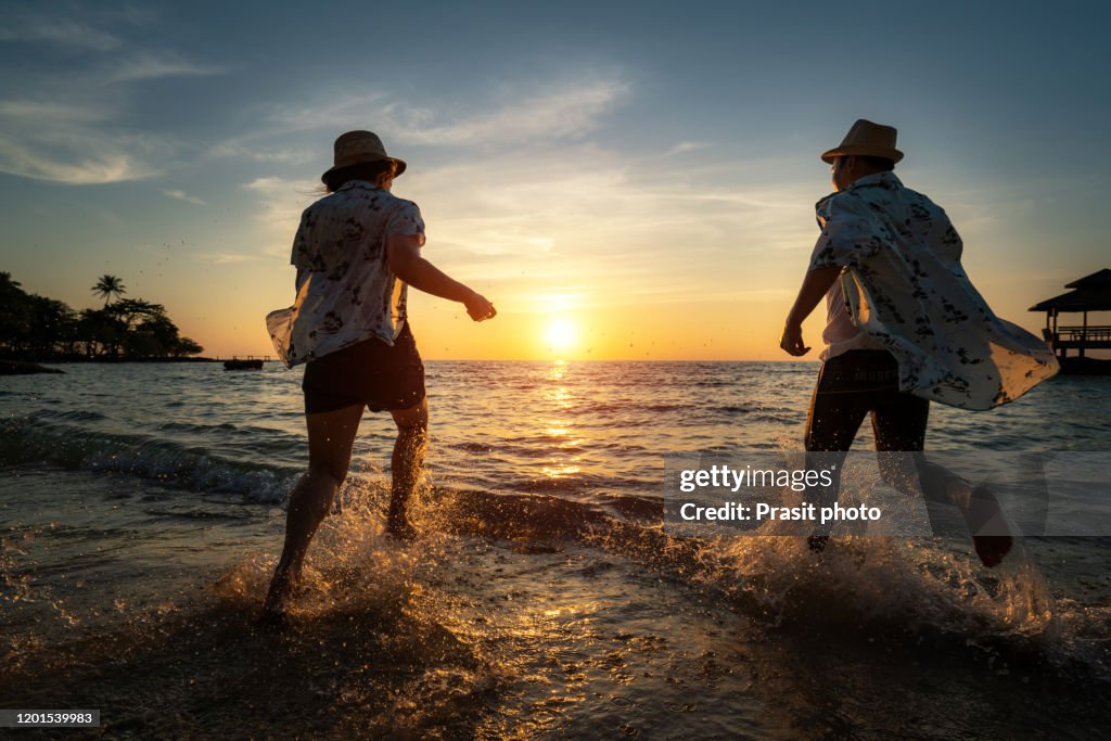 Happy cheerful Asian couple having fun running to the sea together and doing splashes of water on a tropical beach at sunset - concept about romantic vacation, honeymoon.