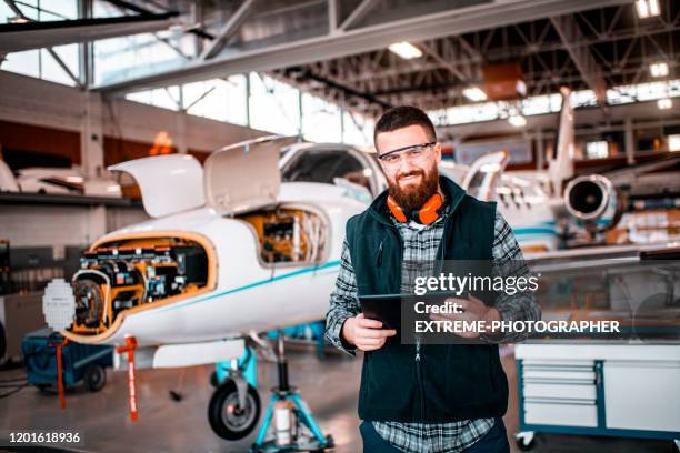 flugzeugmechaniker in einem hangar lächelnd und mit blick auf eine kamera, während sie ein tablet halten und ein flugzeug reparieren und warten - flugzeugmechaniker stock-fotos und bilder