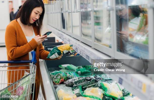 young asian woman scanning barcode on packaging with smart phone in grocery store - gefrierkost stock-fotos und bilder
