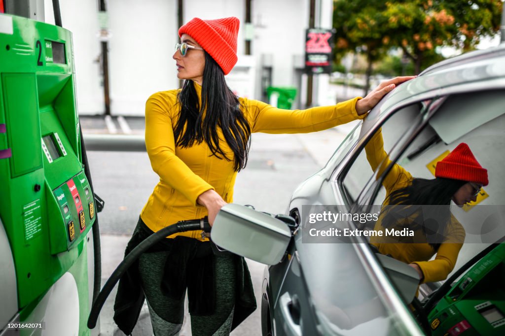 Una mujer moderna y de moda repostando su coche en una gasolinera, mirando a una pantalla de bombeo