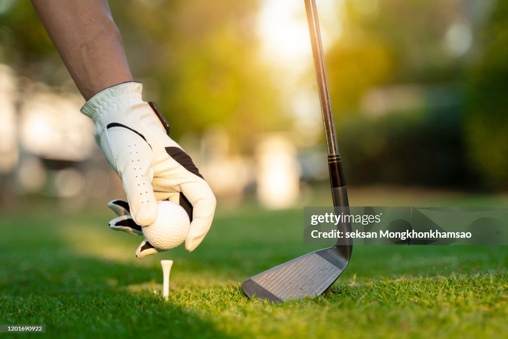 Hand asian woman putting golf ball on tee with club in golf course on evening and sunset time a for healthy sport. Lifestyle Concept