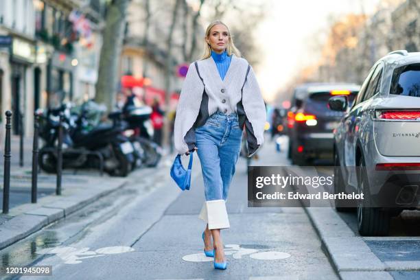 Leonie Hanne wears earrings, a blue turtleneck, a white oversized V-neck knit jacket with grey inserts, a blue Prada handbag, blue crop jeans with...