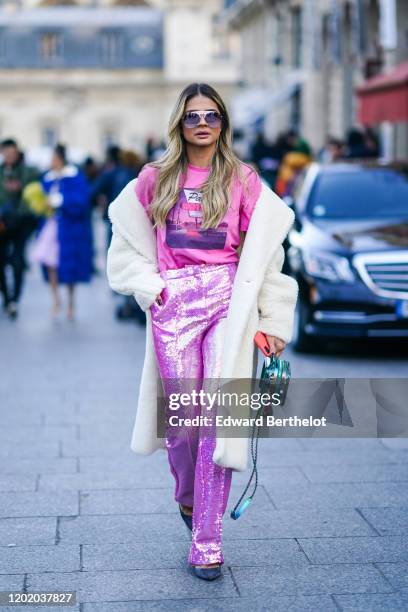 Thassia Naves wears sunglasses, a white fluffy winter long coat, a pink t-shirt with a printed picture, pink/purple glitter shiny sequined flare...