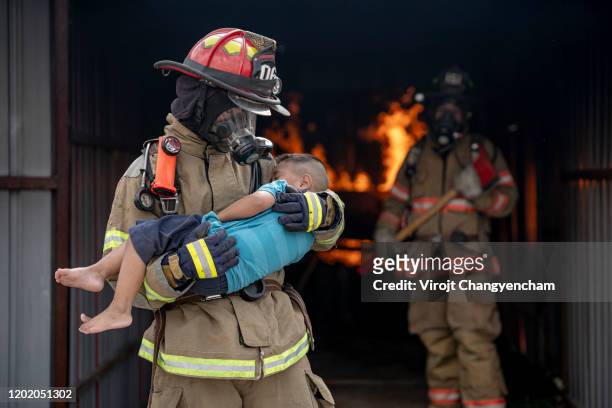 brave fireman of a burning building and holds saved boy in his arms. open fire and one firefighter in the background. - brand advocacy stock-fotos und bilder