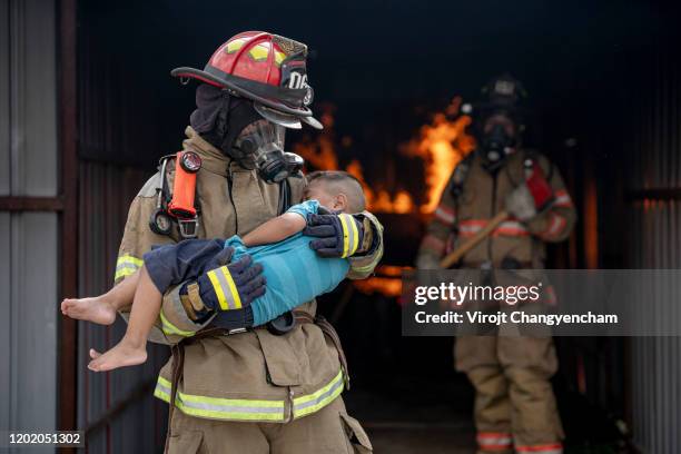 brave fireman of a burning building and holds saved boy in his arms. open fire and one firefighter in the background. - frige bildbanksfoton och bilder