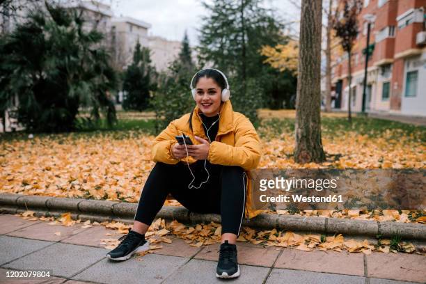 portrait of smiling young woman  listening music with headphones and smartphone in autumn - down coat stock pictures, royalty-free photos & images