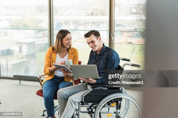 happy young businessman with laptop in wheelchair and businesswoman with tablet in office - gelijkwaardige behandeling stockfoto's en -beelden