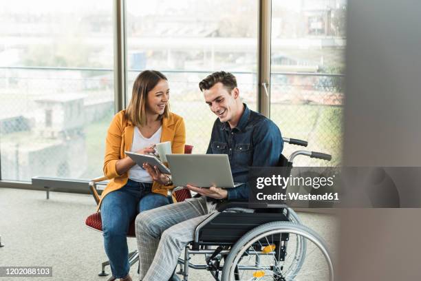 happy young businessman with laptop in wheelchair and businesswoman with tablet in office - young men laughing stock pictures, royalty-free photos & images