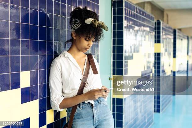 portrait of young woman with curly black hair leaning against tiled wall text messaging - crossbodyväska bildbanksfoton och bilder