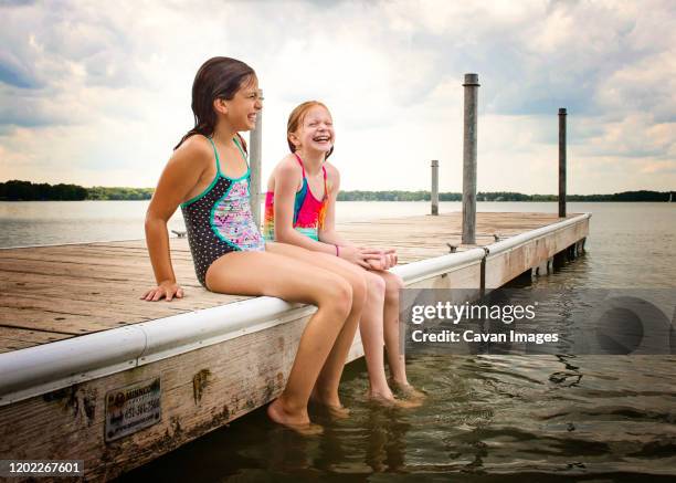 two young girls in swimsuits sitting on a dock by lake laughing - lake auburn stock pictures, royalty-free photos & images