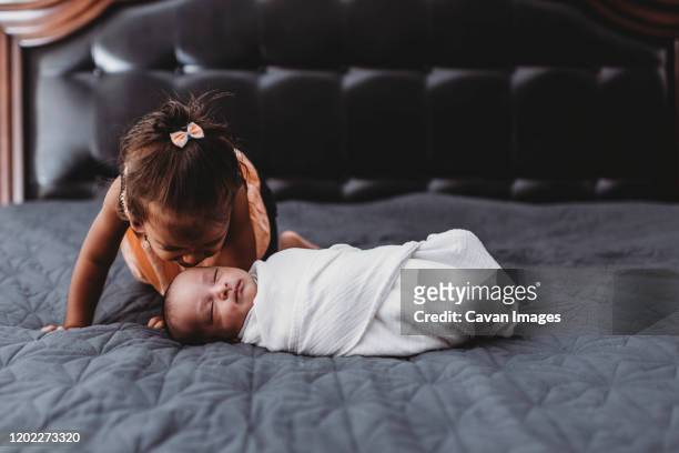 sweet multiracial 2 yr old girl on bed with swaddled infant sibling - babydeken stockfoto's en -beelden