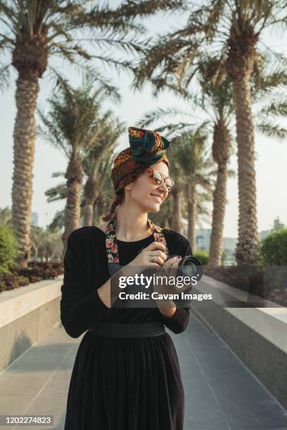 young woman standing in palm alley wearing headwrap and holding camera - doha stock pictures, royalty-free photos & images