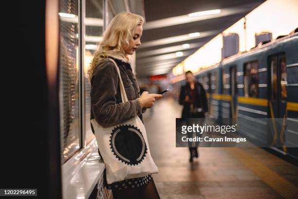 young woman using smart phone while standing at subway station - henkeltasche stock-fotos und bilder