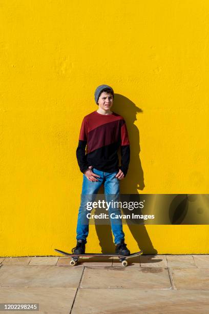 teen with wool cap standing on a skateboard against a yellow wall - hands in pockets foto e immagini stock