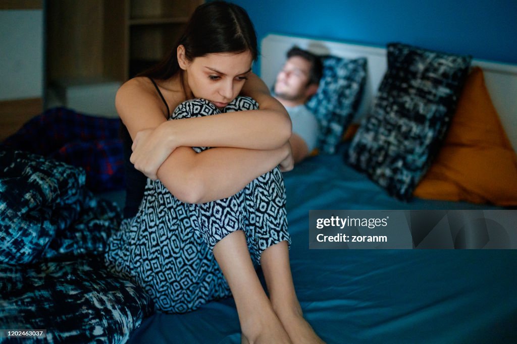 Sad woman sitting on bed with partner in background
