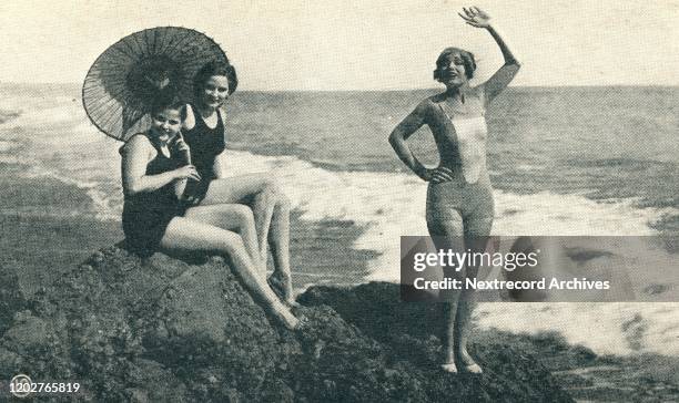 Vintage souvenir postcard published 1920s depicting three movie actresses posing in bathing suits and sun parasol on the beach as promotion for...