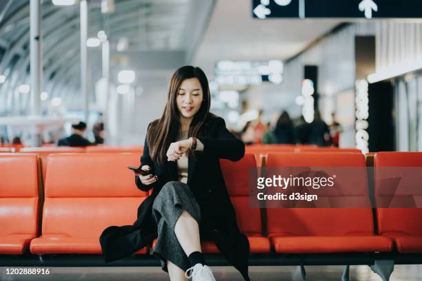 smiling young asian woman holding smartphone on hand and checking time on wristwatch while waiting for her flight in airport lounge - airport waiting stock pictures, royalty-free photos & images