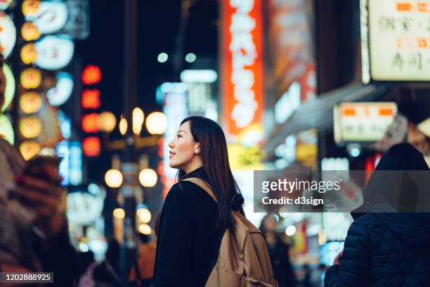 smiling young asian female traveller exploring and strolling along the busy and colourful neon signboard downtown city street at night in osaka, japan - osaka prefecture stock pictures, royalty-free photos & images