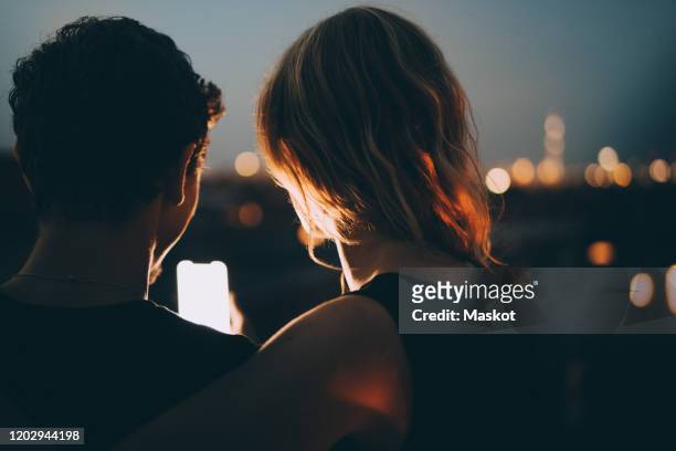 close-up rear view of couple looking at mobile phone while sitting on illuminated terrace in city at dusk - couple in evening clothes stock-fotos und bilder
