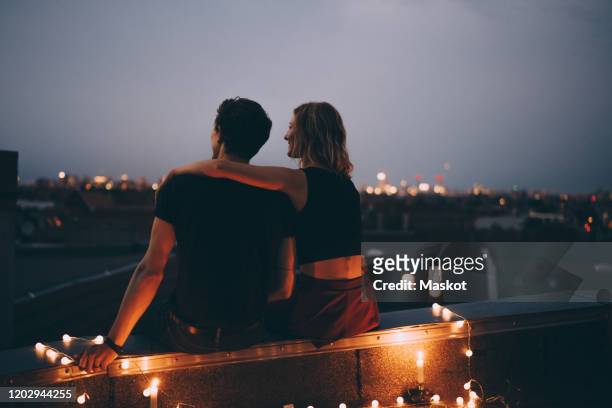 rear view of couple with arm around sitting on illuminated terrace in city against sky - sehen stock-fotos und bilder