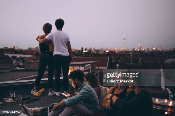 couple looking at city while friends relaxing on terrace during rooftop party - berlin stock-fotos und bilder